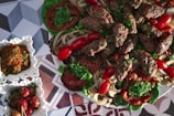 A platter of grilled meat kebabs garnished with fresh parsley, surrounded by grilled vegetables including onions and tomatoes. The dish is accompanied by small square bowls containing a dip or sauce and pickled vegetables, all placed on a geometric patterned surface.
