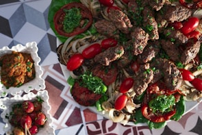 A platter of grilled meat kebabs garnished with fresh parsley, surrounded by grilled vegetables including onions and tomatoes. The dish is accompanied by small square bowls containing a dip or sauce and pickled vegetables, all placed on a geometric patterned surface.