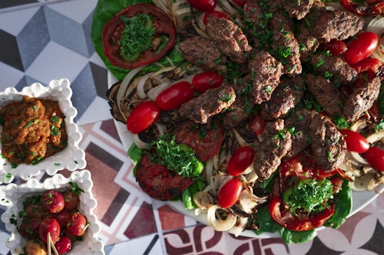 A platter of grilled meat kebabs garnished with fresh parsley, surrounded by grilled vegetables including onions and tomatoes. The dish is accompanied by small square bowls containing a dip or sauce and pickled vegetables, all placed on a geometric patterned surface.