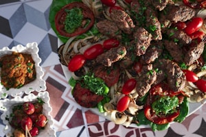 A platter of grilled meat kebabs garnished with fresh parsley, surrounded by grilled vegetables including onions and tomatoes. The dish is accompanied by small square bowls containing a dip or sauce and pickled vegetables, all placed on a geometric patterned surface.