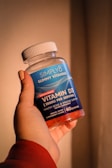 A hand holding a bottle of Simply8 Gummy Vitamins, specifically Vitamin D3 with a dosage of 2000 IU per serving. The bottle is blue with a white lid and contains 60 gummies with orange, lemon, and strawberry flavor.