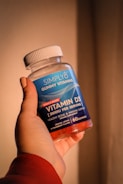 A hand holding a bottle of Simply8 Gummy Vitamins, specifically Vitamin D3 with a dosage of 2000 IU per serving. The bottle is blue with a white lid and contains 60 gummies with orange, lemon, and strawberry flavor.