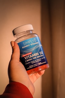 A hand holding a bottle of Simply8 Gummy Vitamins, specifically Vitamin D3 with a dosage of 2000 IU per serving. The bottle is blue with a white lid and contains 60 gummies with orange, lemon, and strawberry flavor.