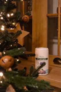 A container of pet ear wipes is placed on a wooden surface. In the foreground, Christmas tree branches are adorned with yellow lights and wooden star-shaped ornaments, creating a cozy festive atmosphere.