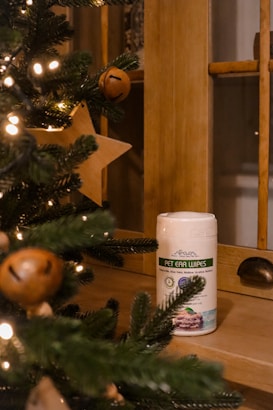 A container of pet ear wipes is placed on a wooden surface. In the foreground, Christmas tree branches are adorned with yellow lights and wooden star-shaped ornaments, creating a cozy festive atmosphere.