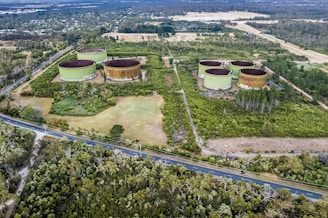 An aerial view of a landscaped area with several large, circular, green industrial tanks surrounded by greenery and trees. There are roads running through and around the area, with dense forested regions nearby.