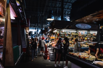 A bustling indoor market with numerous stalls selling a variety of products. People are shopping and interacting, creating a lively atmosphere. The stalls are packed with colorful goods, including cans and fresh produce.