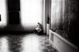 An elderly person sits quietly on a chair by a large window, holding a newspaper. The room is softly lit with natural light streaming through vertical blinds, revealing a polished wooden floor. Large paintings hang on the walls, adding an artistic atmosphere to the setting.