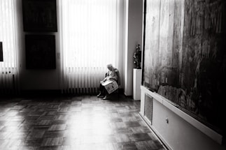 An elderly person sits quietly on a chair by a large window, holding a newspaper. The room is softly lit with natural light streaming through vertical blinds, revealing a polished wooden floor. Large paintings hang on the walls, adding an artistic atmosphere to the setting.