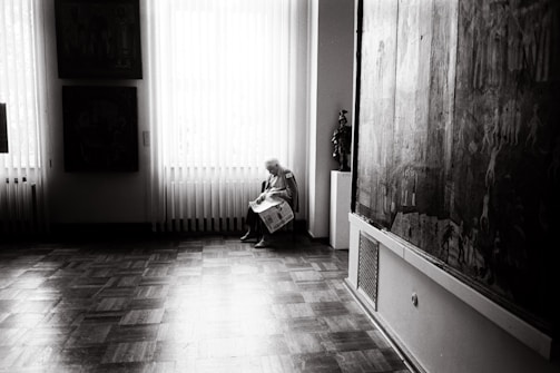 An elderly person sits quietly on a chair by a large window, holding a newspaper. The room is softly lit with natural light streaming through vertical blinds, revealing a polished wooden floor. Large paintings hang on the walls, adding an artistic atmosphere to the setting.