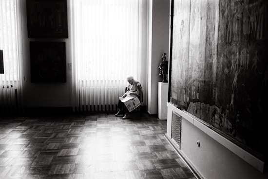 An elderly person sits quietly on a chair by a large window, holding a newspaper. The room is softly lit with natural light streaming through vertical blinds, revealing a polished wooden floor. Large paintings hang on the walls, adding an artistic atmosphere to the setting.