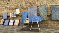 An assortment of colorful artworks displayed against a rustic stone wall. Several fabric pieces with intricate, geometric designs in shades of blue, along with framed paintings are arranged on the stone pavement. Among these, a wooden chair and easel are partially visible, adding to the artistic ambiance.