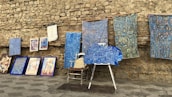 An assortment of colorful artworks displayed against a rustic stone wall. Several fabric pieces with intricate, geometric designs in shades of blue, along with framed paintings are arranged on the stone pavement. Among these, a wooden chair and easel are partially visible, adding to the artistic ambiance.