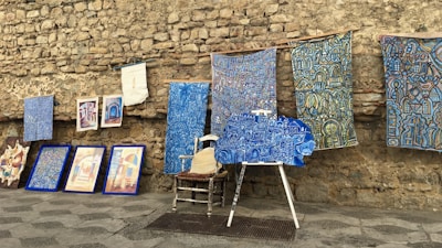 An assortment of colorful artworks displayed against a rustic stone wall. Several fabric pieces with intricate, geometric designs in shades of blue, along with framed paintings are arranged on the stone pavement. Among these, a wooden chair and easel are partially visible, adding to the artistic ambiance.