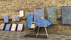 An assortment of colorful artworks displayed against a rustic stone wall. Several fabric pieces with intricate, geometric designs in shades of blue, along with framed paintings are arranged on the stone pavement. Among these, a wooden chair and easel are partially visible, adding to the artistic ambiance.