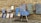 An assortment of colorful artworks displayed against a rustic stone wall. Several fabric pieces with intricate, geometric designs in shades of blue, along with framed paintings are arranged on the stone pavement. Among these, a wooden chair and easel are partially visible, adding to the artistic ambiance.