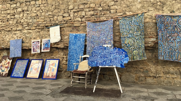 An assortment of colorful artworks displayed against a rustic stone wall. Several fabric pieces with intricate, geometric designs in shades of blue, along with framed paintings are arranged on the stone pavement. Among these, a wooden chair and easel are partially visible, adding to the artistic ambiance.