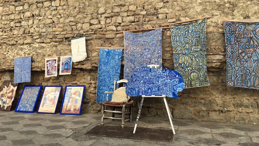 An assortment of colorful artworks displayed against a rustic stone wall. Several fabric pieces with intricate, geometric designs in shades of blue, along with framed paintings are arranged on the stone pavement. Among these, a wooden chair and easel are partially visible, adding to the artistic ambiance.
