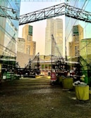 A modern urban scene with tall skyscrapers, reflective glass façades, and architectural elements creating geometric patterns. The ground level contains potted plants and a sign for stores like Superdry, suggesting a shopping or commercial area.