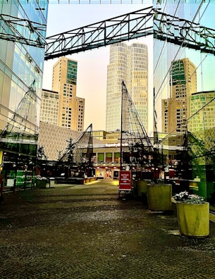 A modern urban scene with tall skyscrapers, reflective glass façades, and architectural elements creating geometric patterns. The ground level contains potted plants and a sign for stores like Superdry, suggesting a shopping or commercial area.