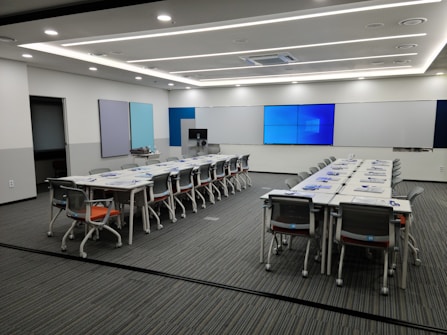 A spacious and modern conference room with a long rectangular table set up with chairs around it. The walls are adorned with multiple screens and minimalistic decor. The lighting is bright and evenly distributed across the room, giving it a professional atmosphere.