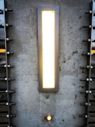 A rectangular light fixture set into a textured concrete wall. The light emits a warm glow, surrounded by a metallic frame. There are symmetrical metal bolts fixed into the wall on either side of the light. Below the main light, there is a smaller circular light embedded in the wall.