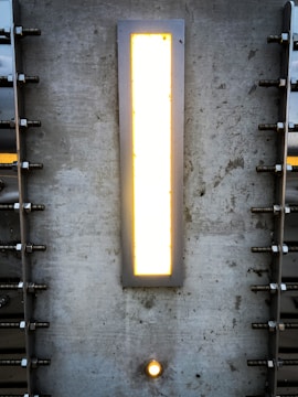A rectangular light fixture set into a textured concrete wall. The light emits a warm glow, surrounded by a metallic frame. There are symmetrical metal bolts fixed into the wall on either side of the light. Below the main light, there is a smaller circular light embedded in the wall.