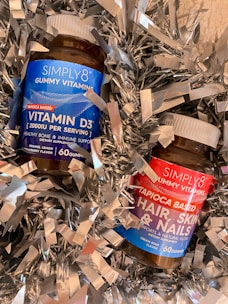 Two bottles of Simply8 gummy vitamins are surrounded by metallic silver decorative shreds. One bottle is labeled as Vitamin D3 with a blue label, while the other is for hair, skin, and nails with a red label. Both bottles have white caps.
