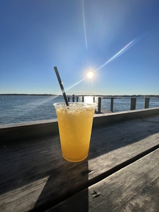 A vibrant yellow drink in a clear plastic cup with ice and a black straw sits on a wooden surface. The background features a calm body of water and a bright sun in a clear blue sky, creating a serene and refreshing atmosphere.