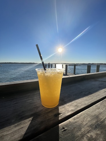 A vibrant yellow drink in a clear plastic cup with ice and a black straw sits on a wooden surface. The background features a calm body of water and a bright sun in a clear blue sky, creating a serene and refreshing atmosphere.