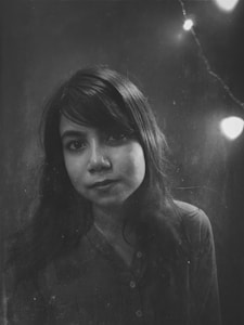 A black and white portrait of a woman with long dark hair, looking directly at the camera. The background is dark and faded, featuring a subtle string of lights in the upper right corner adding a soft glowing effect.