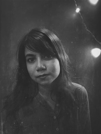 A black and white portrait of a woman with long dark hair, looking directly at the camera. The background is dark and faded, featuring a subtle string of lights in the upper right corner adding a soft glowing effect.