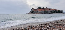 A picturesque island with charming old buildings featuring red-tiled roofs is surrounded by the sea. The shoreline is lined with a pebble beach, and gentle waves approach the shore. The sky is overcast, giving the scene a calm and serene atmosphere.
