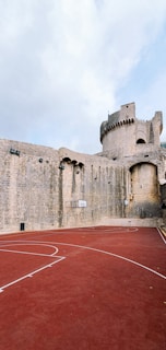 A basketball court with a red surface is situated next to an ancient stone fortress. The fortress features tall stone walls and a tower-like structure, suggesting historical architecture. The basketball court's lines are marked in white, and two hoops are attached to the fortress walls.