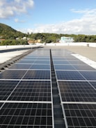 A large array of solar panels is installed on a flat rooftop under a bright blue sky with scattered clouds. In the distance, there is a lush green hillside with trees and several residential buildings with colorful rooftops.