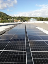 A large array of solar panels is installed on a flat rooftop under a bright blue sky with scattered clouds. In the distance, there is a lush green hillside with trees and several residential buildings with colorful rooftops.