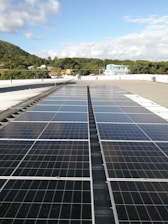 A large array of solar panels is installed on a flat rooftop under a bright blue sky with scattered clouds. In the distance, there is a lush green hillside with trees and several residential buildings with colorful rooftops.
