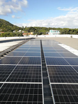 A large array of solar panels is installed on a flat rooftop under a bright blue sky with scattered clouds. In the distance, there is a lush green hillside with trees and several residential buildings with colorful rooftops.