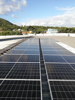 A large array of solar panels is installed on a flat rooftop under a bright blue sky with scattered clouds. In the distance, there is a lush green hillside with trees and several residential buildings with colorful rooftops.