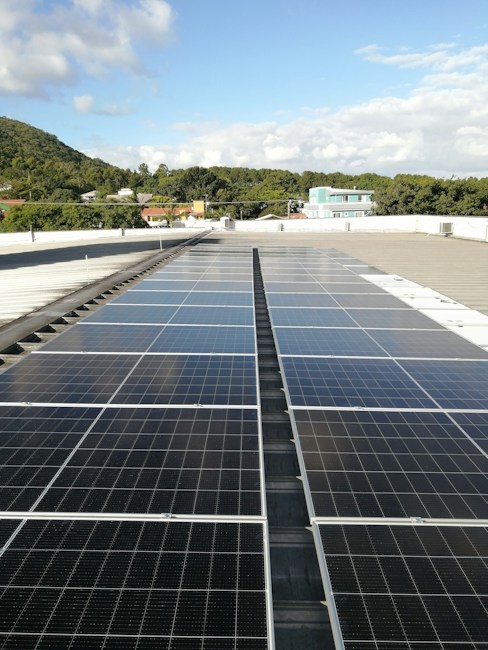 A large array of solar panels is installed on a flat rooftop under a bright blue sky with scattered clouds. In the distance, there is a lush green hillside with trees and several residential buildings with colorful rooftops.
