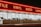 A storefront with large windows illuminated from within, featuring a bold, red neon sign displaying words like 'TILE', 'VINYL', 'WOOD', and 'LAMIN'. The exterior is dimly lit, creating a stark contrast between the neon light and the night sky.
