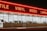 A storefront with large windows illuminated from within, featuring a bold, red neon sign displaying words like 'TILE', 'VINYL', 'WOOD', and 'LAMIN'. The exterior is dimly lit, creating a stark contrast between the neon light and the night sky.