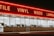 A storefront with large windows illuminated from within, featuring a bold, red neon sign displaying words like 'TILE', 'VINYL', 'WOOD', and 'LAMIN'. The exterior is dimly lit, creating a stark contrast between the neon light and the night sky.