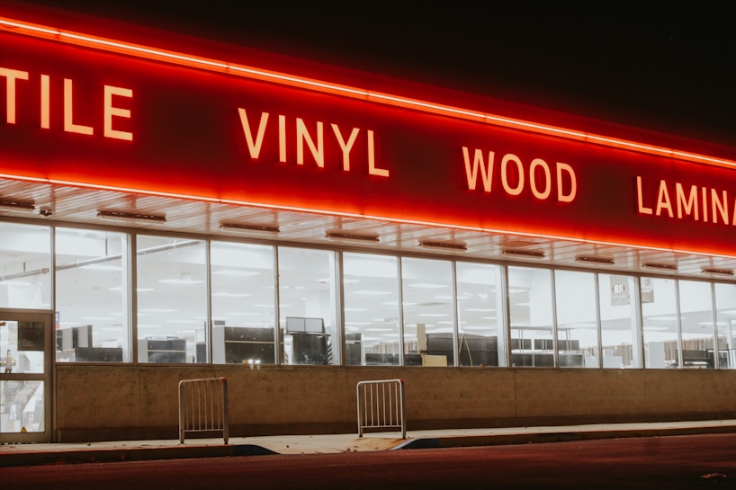 A storefront with large windows illuminated from within, featuring a bold, red neon sign displaying words like 'TILE', 'VINYL', 'WOOD', and 'LAMIN'. The exterior is dimly lit, creating a stark contrast between the neon light and the night sky.
