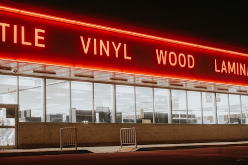 A storefront with large windows illuminated from within, featuring a bold, red neon sign displaying words like 'TILE', 'VINYL', 'WOOD', and 'LAMIN'. The exterior is dimly lit, creating a stark contrast between the neon light and the night sky.