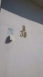 A white wall features a doorbell with a sign next to it that says 'BELL' and an arrow pointing to the push button. Above, there are metallic house numbers and a letter 'B 38'. The shadow of the lettering is visible on the wall.