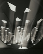 An expansive, abstract architectural space with large circular and triangular openings in the ceiling allowing beams of light to stream down. The atmosphere is ethereal, as light interacts with the geometric patterns and shadows, creating a sense of depth and scale. A solitary figure stands at the center, adding a human element to the surreal environment.