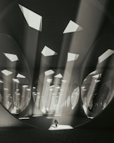An expansive, abstract architectural space with large circular and triangular openings in the ceiling allowing beams of light to stream down. The atmosphere is ethereal, as light interacts with the geometric patterns and shadows, creating a sense of depth and scale. A solitary figure stands at the center, adding a human element to the surreal environment.