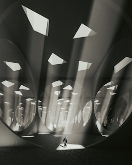 An expansive, abstract architectural space with large circular and triangular openings in the ceiling allowing beams of light to stream down. The atmosphere is ethereal, as light interacts with the geometric patterns and shadows, creating a sense of depth and scale. A solitary figure stands at the center, adding a human element to the surreal environment.