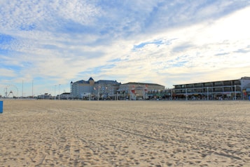 A wide sandy beach stretches into the distance, leading to a boardwalk lined with buildings. A Ferris wheel is visible on the left side near the horizon, among other amusement structures. The sky is partly cloudy, with soft wisps of white and light blue.